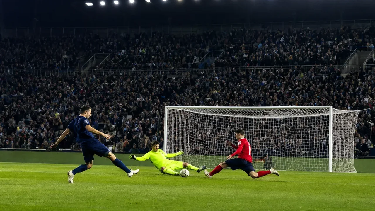 A striker in navy shooting as a defender slides in and the goalkeeper dives under floodlights